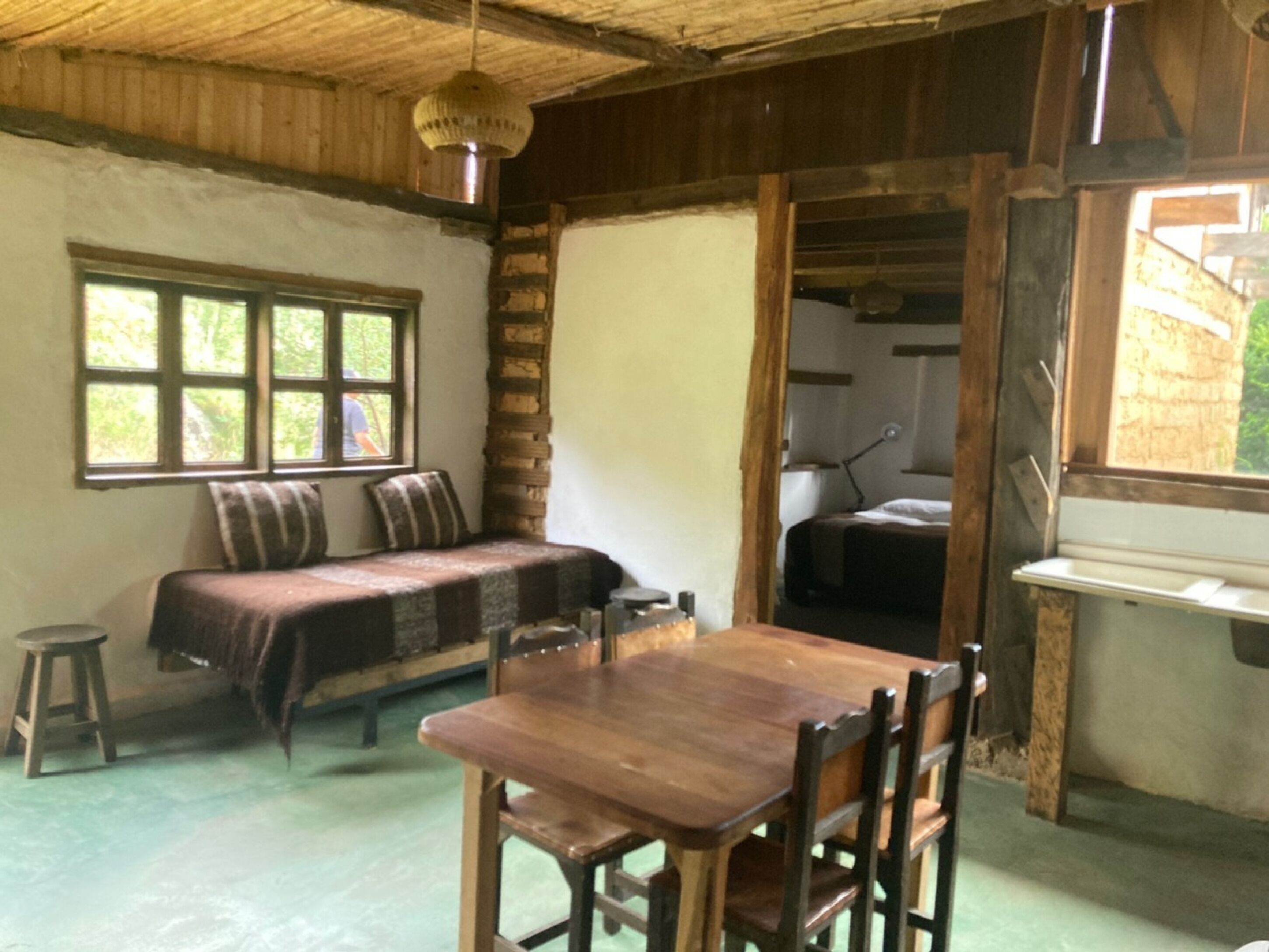 A rustic room with a wooden table and chairs, a bench with pillows, a window, and a door leading to another room.