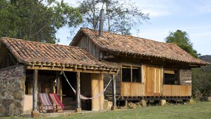 Una cabaña rústica de madera con techo de tejas, un porche con dos sillas a rayas y una hamaca.