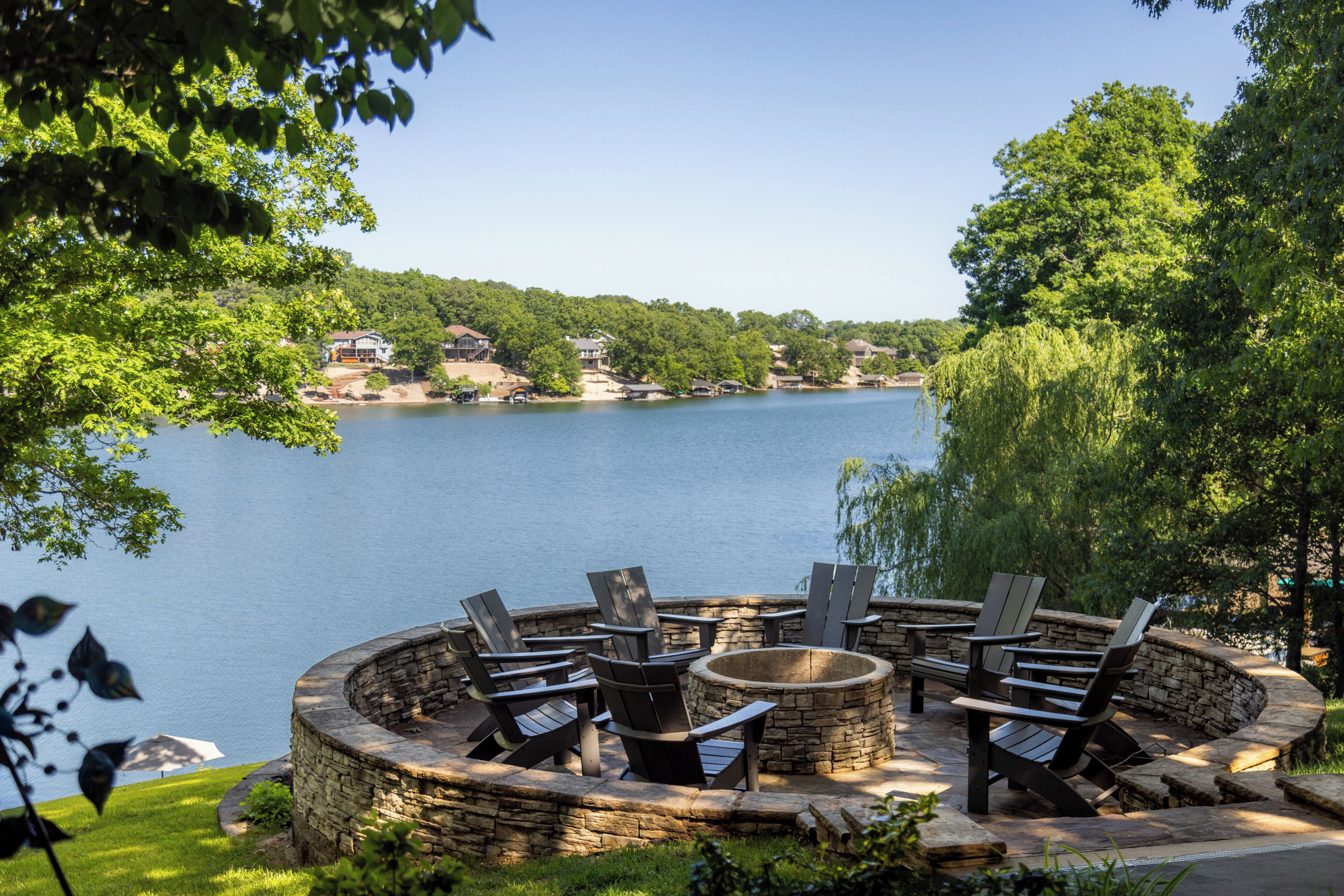 A circular stone fire pit with Adirondack chairs around it, overlooking a lake with houses in the background.