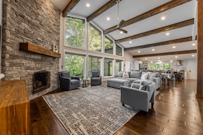 A spacious living area with a stone fireplace, large windows, and a high ceiling with exposed beams.