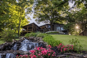 A modern house with a waterfall, surrounded by lush greenery and flowering plants.