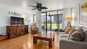 A spacious living room with a wooden coffee table, a grey sofa with patterned cushions, a television on a wooden cabinet, and a large window offering a view of the outdoors. - Spacious 1 Bedroom Condo at Turtle Bay (Kahuku)