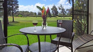 A round table with two chairs, a vase with flowers, and two wine glasses on a balcony overlooking a golf course. - Spacious 1 Bedroom Condo at Turtle Bay (Kahuku)