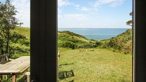 Blick aus einem Gebäude auf ein grünes Feld mit einer Holzbank und einem Tisch, sowie ein Gewässer in der Ferne.