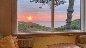 Ein Zimmer mit großem Fenster und Blick auf einen Sonnenuntergang über einer hügeligen Landschaft. Es gibt einen Holztisch und zwei Polsterstühle mit Kissen.