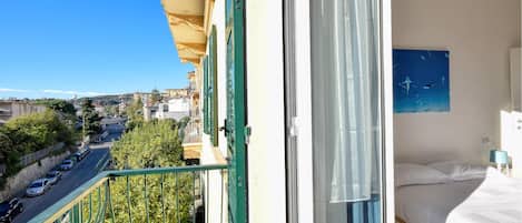 A balcony with a view of a street and buildings, a bed with a white mattress and pillows, and a painting of sharks on the wall.