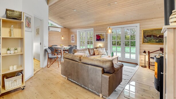 A cozy living room with a wooden ceiling, a brown leather sofa, a dining area with chairs, and a kitchen area with a stove. - 3 bedroom gorgeous home in Slagelse (Slagelse)