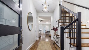 A modern interior with a wooden staircase, a kitchen in the background, and a hallway leading to a dining area. - Spacious 4BR Retreat, Sleeps 10, Backyard + Near Dallas (Dallas)