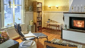 Un salon chaleureux avec une cheminée, une bibliothèque, une fenêtre donnant sur un paysage enneigé et un fauteuil à motifs.