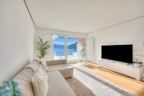 A modern living room with a sofa, a flat-screen TV, a plant, and a view of the outdoors. - Honeymoon With Panoramic Lake-view (Lugano)