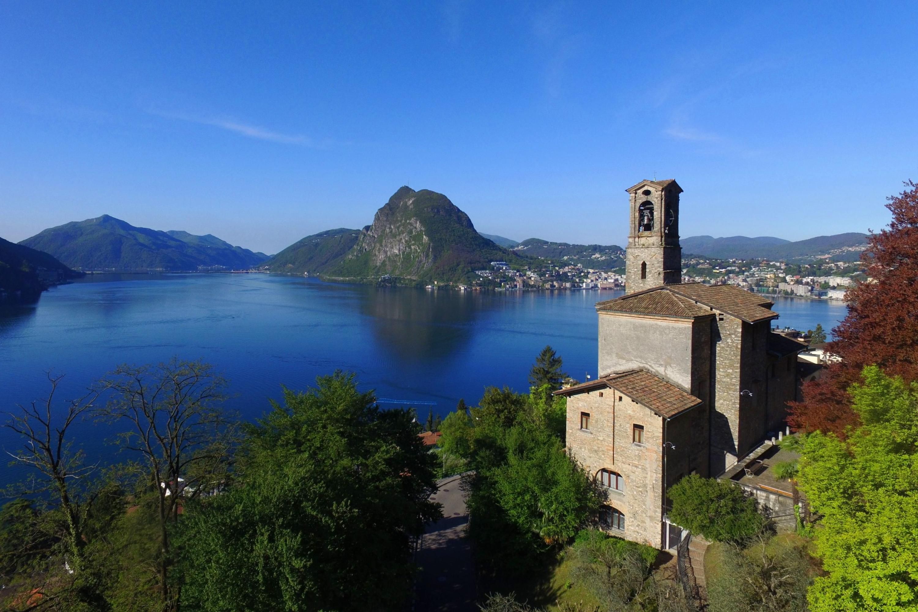 A stone building with a bell tower, surrounded by trees and overlooking a calm lake with mountains in the background.