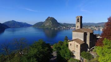 A stone building with a bell tower, surrounded by trees and overlooking a calm lake with mountains in the background.