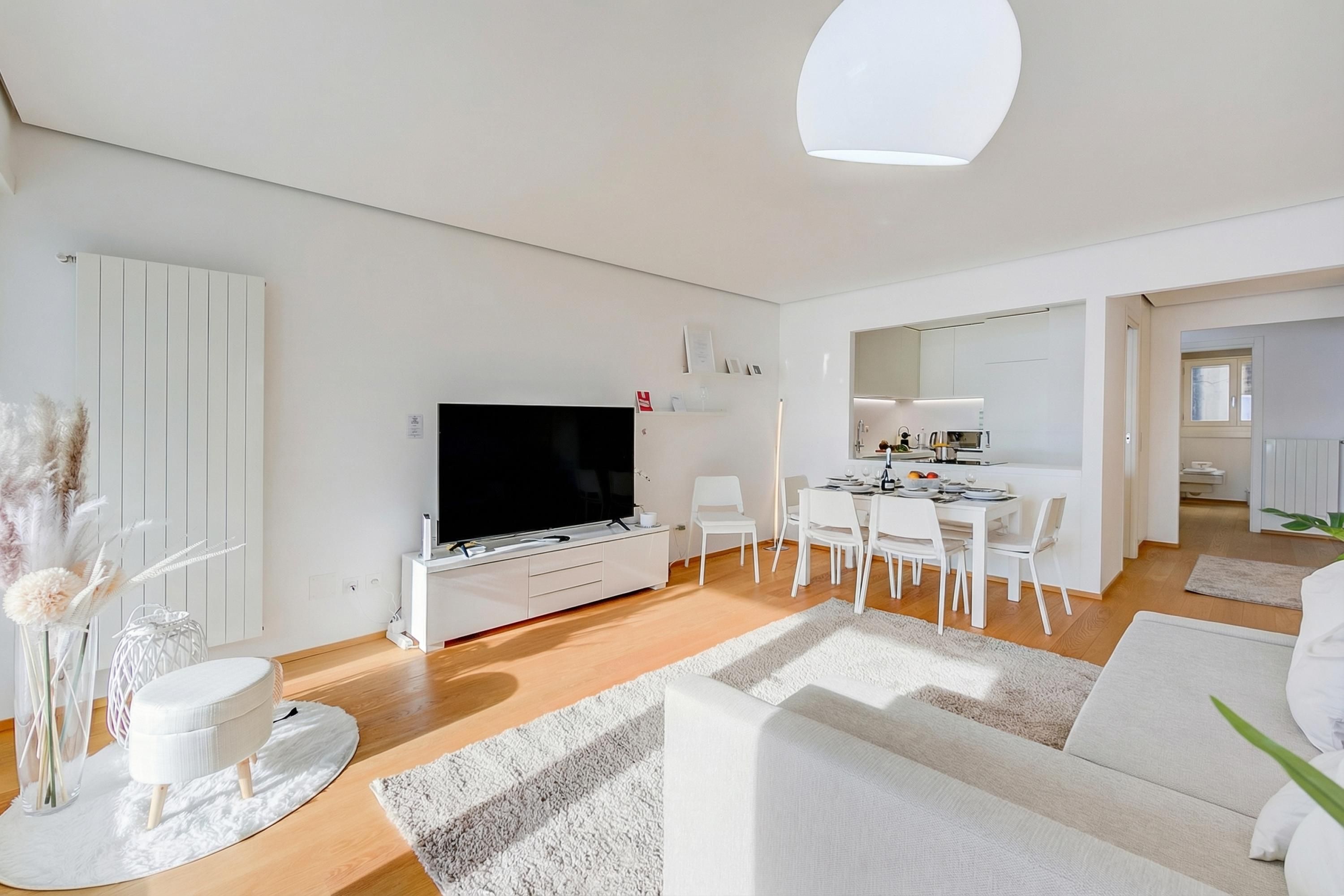 A modern living room with a flat-screen TV, white furniture, and a dining area.
