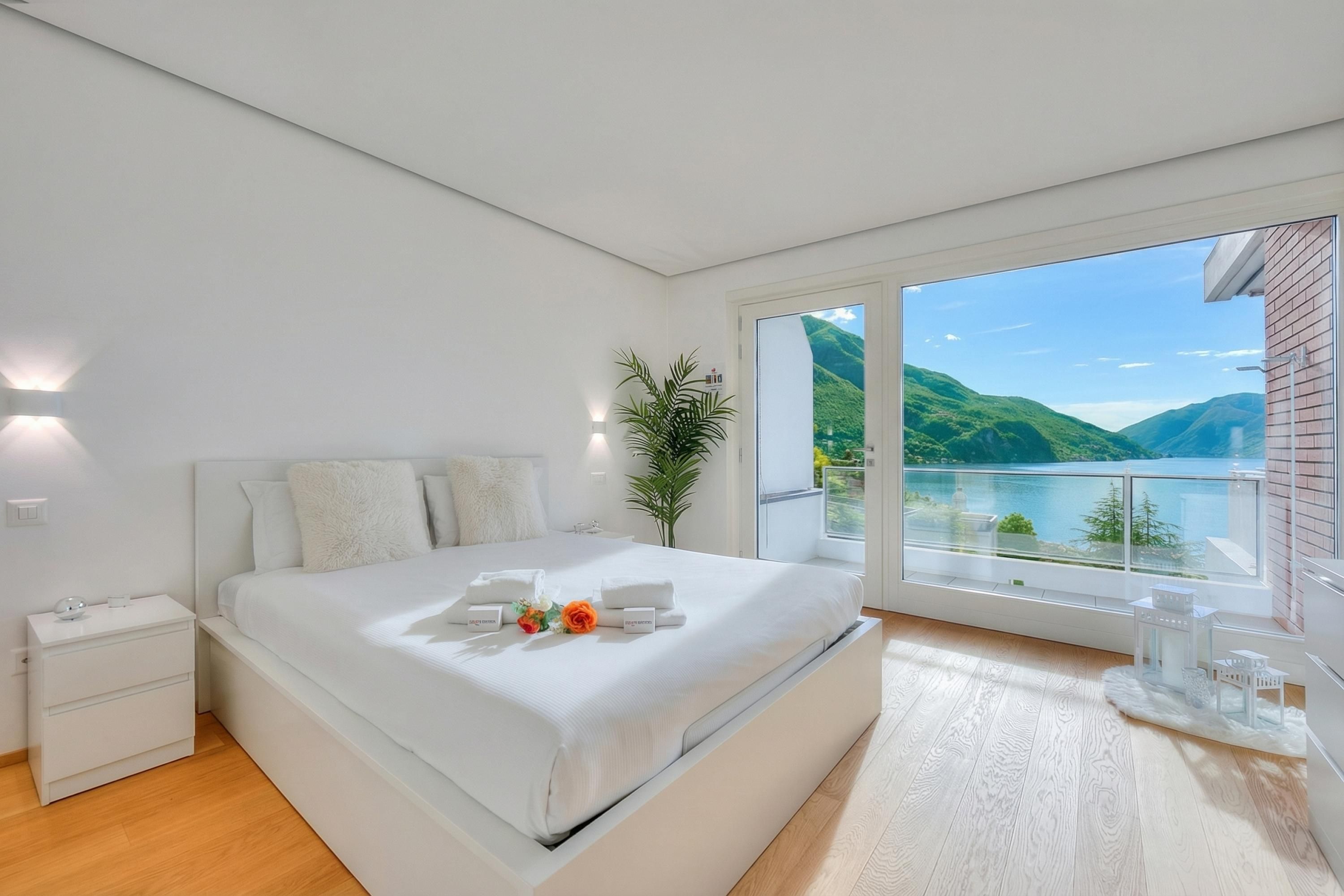A modern bedroom with a large bed, a nightstand, a potted plant, and a view of a lake.