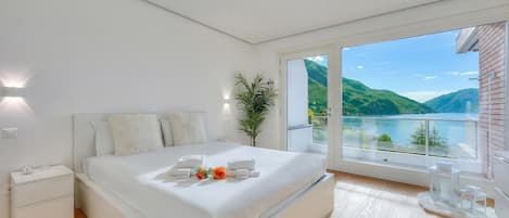 A modern bedroom with a large bed, a nightstand, a potted plant, and a view of a lake.
