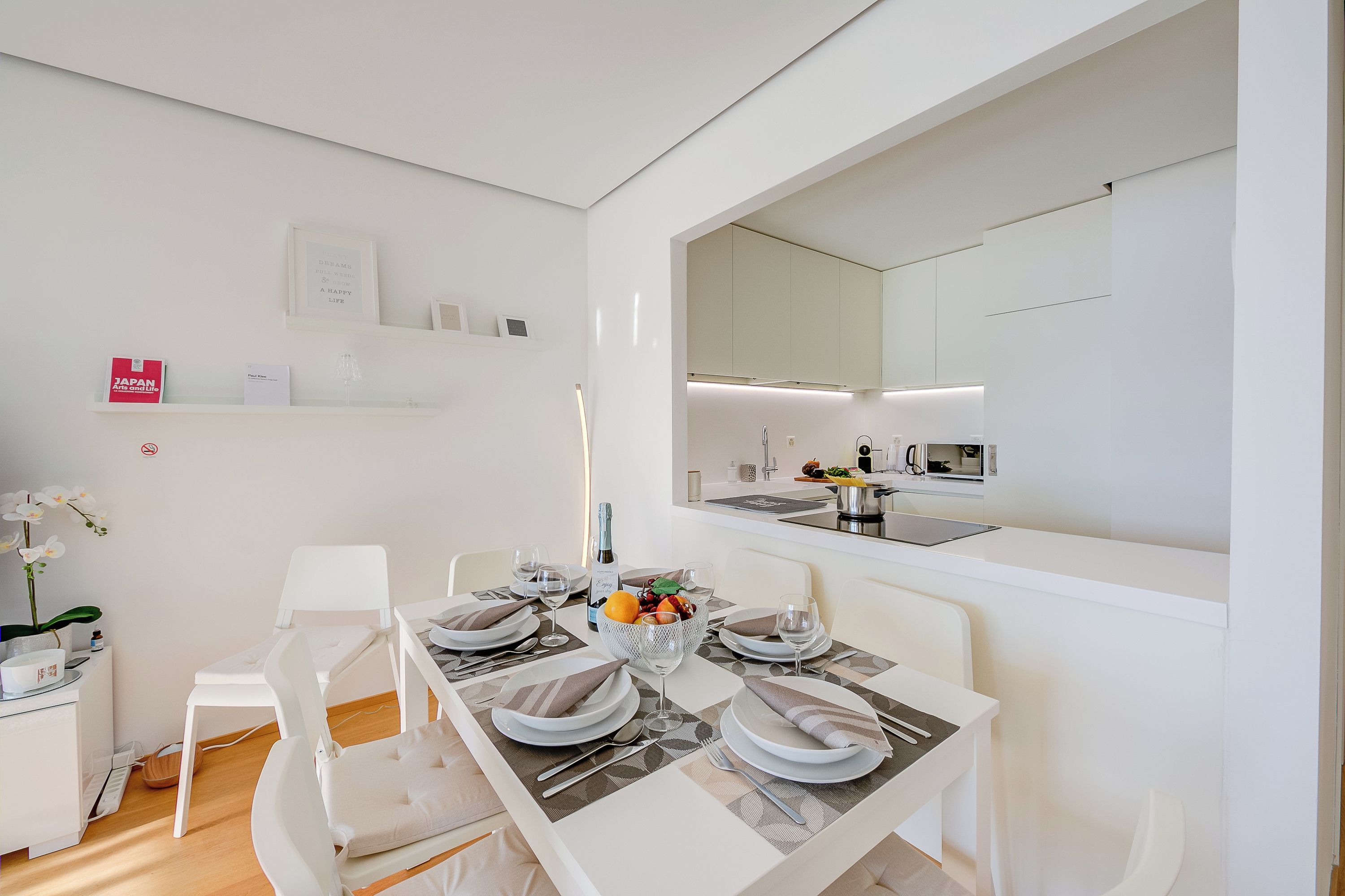 A modern dining area with a white table set for four, a white chair, a small white shelf with books, and a kitchen visible through an open door.