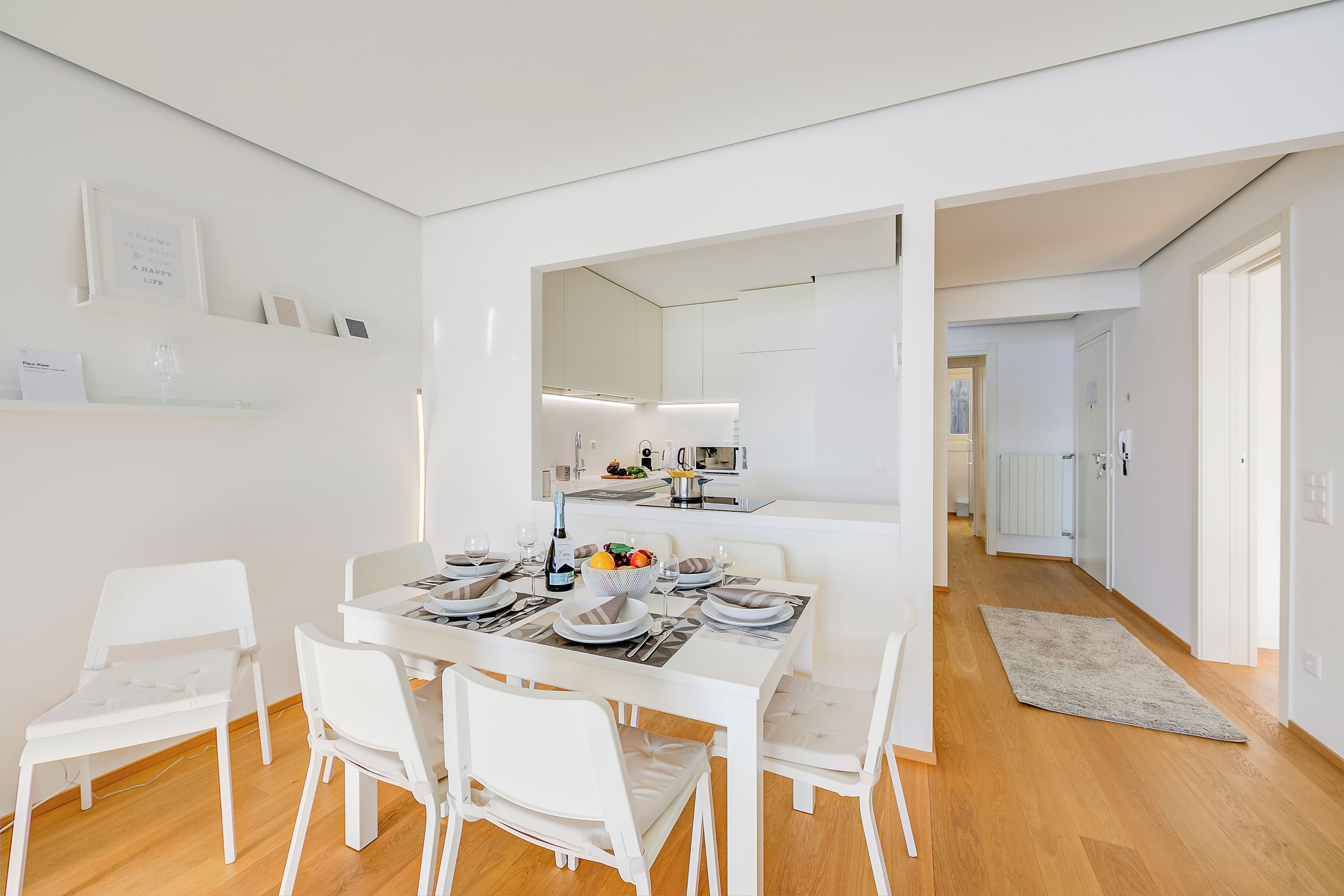 A modern dining area with a white table set for four, surrounded by white chairs. A large mirror reflects the room, and a kitchen is visible in the background.