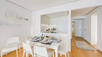 A modern dining area with a white table set for four, surrounded by white chairs. A large mirror reflects the room, and a kitchen is visible in the background.