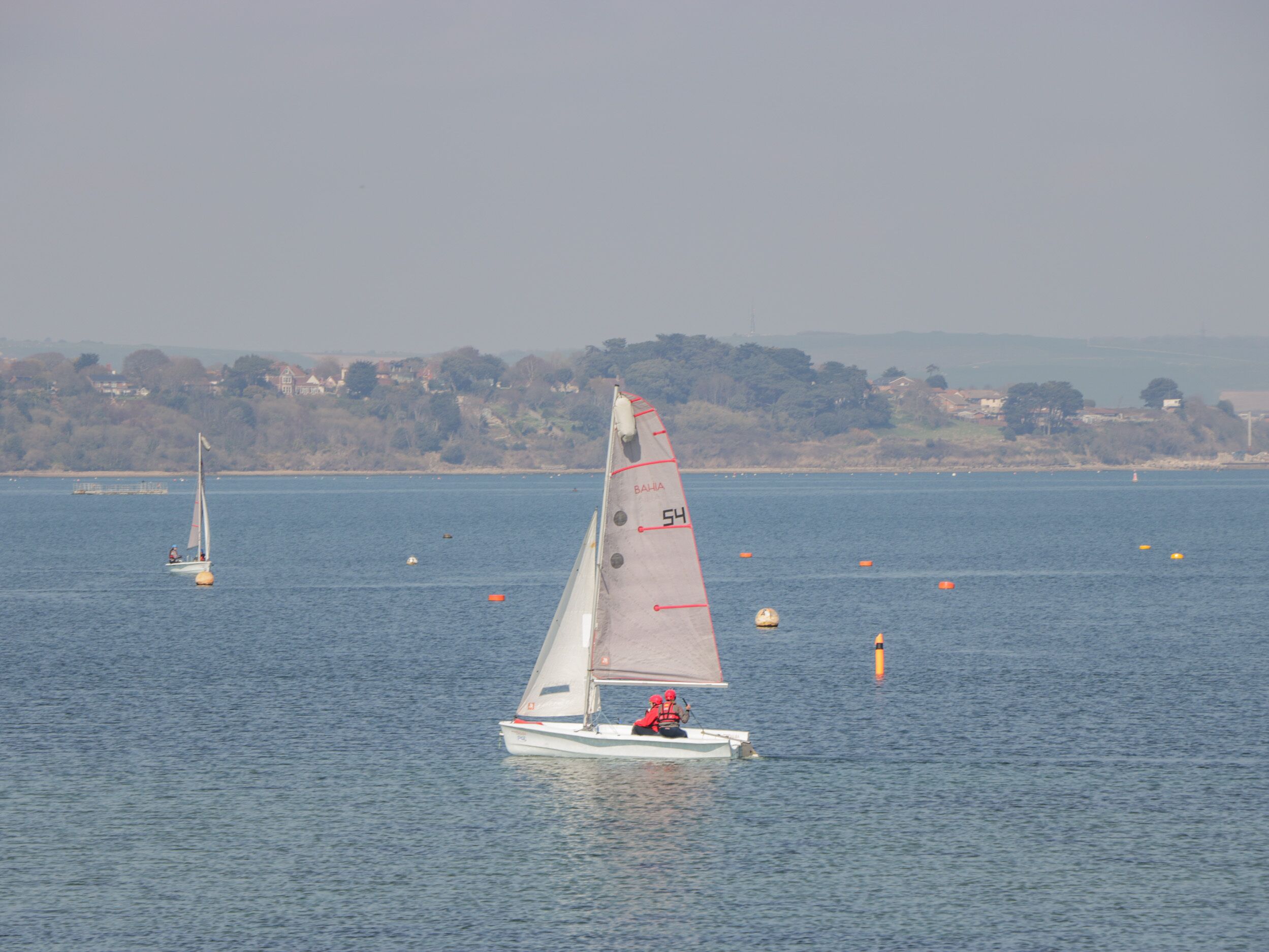 A sailboat on a calm body of water with buoys and a distant shoreline.