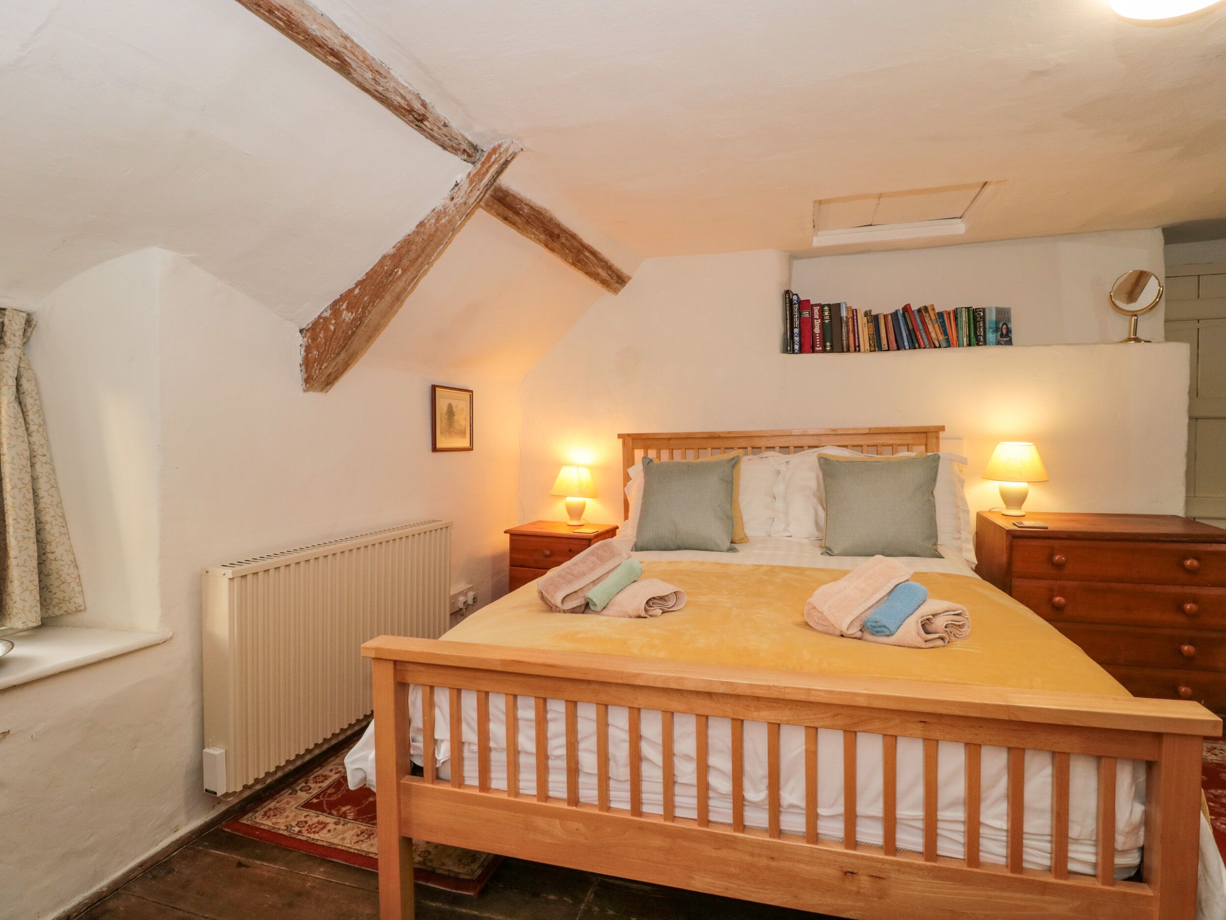 A bedroom with a wooden bed, two bedside tables with lamps, a radiator, a bookshelf, and a window with curtains.