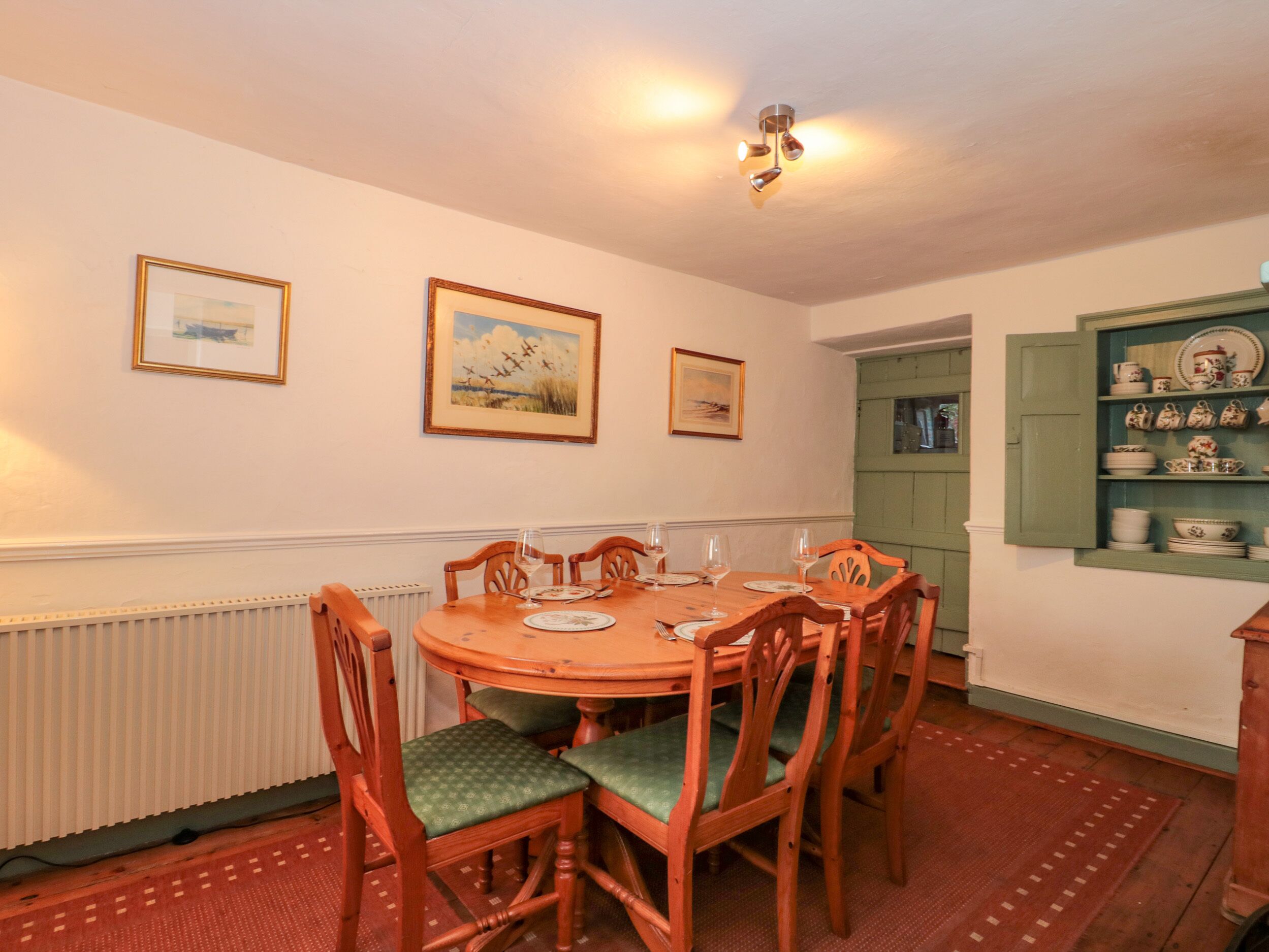 A dining room with a round wooden table, six chairs, and a green patterned rug. There are three framed pictures on the wall, and a green cupboard with dishes.