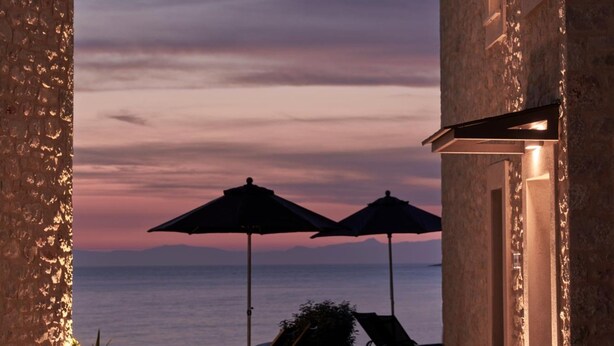 Un edificio de piedra con un patio empedrado, dos sombrillas y vistas al mar al atardecer.
