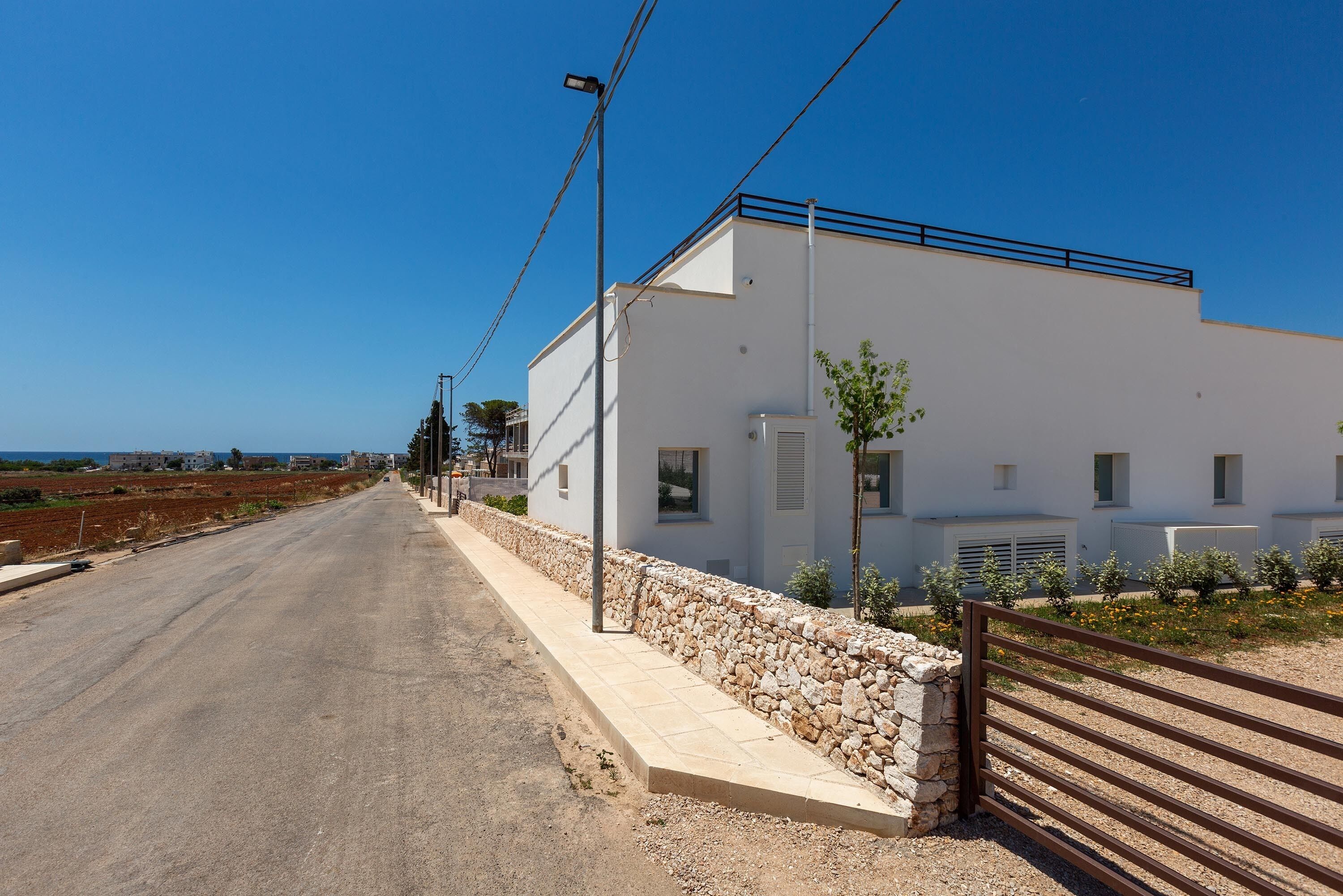 Un edificio moderno con un muro de piedra y una valla metálica, situado al lado de una carretera bajo un cielo despejado.