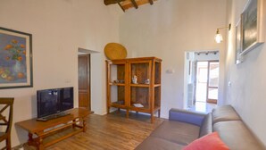 Ein Wohnzimmer mit Holzboden, einem Fernseher auf einem Holztisch, einem Holzschrank mit Glastüren, einer Couch mit Kissen und einem Gemälde an der Wand.