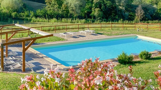 Ein Schwimmbecken mit Holzterrasse und Liegestühlen, umgeben von einer Umzäunung und üppiger Bepflanzung.