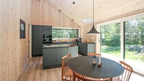 A modern kitchen with wooden walls, a central island, and a dining area with a round table and wooden chairs. - 2 bedroom cozy home in Samsø (Samsø)