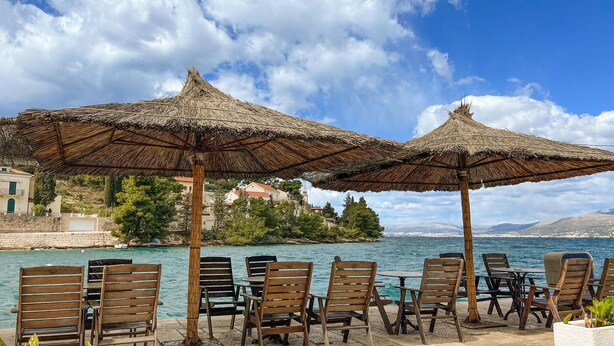 Une terrasse en bord de mer, avec des chaises en bois et des parasols en chaume, offrant une vue sur une mer calme et une ville côtière.