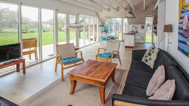 Een ruime zithoek met een houten salontafel, twee fauteuils, een bank en een televisie op een meubel. Grote ramen bieden uitzicht op het omliggende landschap.