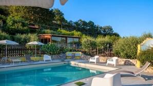 Piscina con sillas de descanso y sombrillas, rodeada de follaje y una estructura de madera.