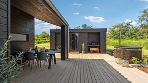 Een modern terras buiten met een houten terras, een eethoek met stoelen en een tafel, een jacuzzi en een klein bijgebouw met een barbecue.