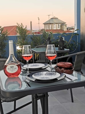 A table set with wine glasses, a bottle of Mateus wine, plates, and a basket of food on a balcony with a view of buildings and plants.