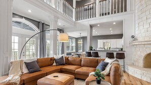 A spacious living room with a large sectional sofa, a fireplace, and a kitchen in the background. - Pet-Friendly! Pool/Spa, Views, Arcade, EV Charger (Wimberley)