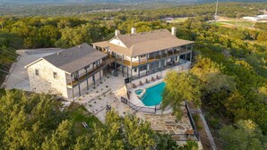 A large two-story house with a swimming pool, surrounded by trees and a hilly landscape. - Pet-Friendly! Pool/Spa, Views, Arcade, EV Charger (Wimberley)