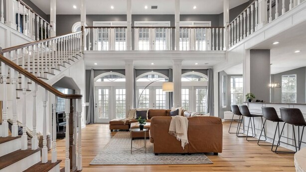 A spacious living area with a high ceiling, a staircase with white railings, and a seating area with a brown sofa and a coffee table. - Pet-Friendly! Pool/Spa, Views, Arcade, EV Charger (Wimberley)