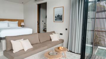 Quarto de hotel moderno com uma cama grande, um sofá, duas mesinhas de apoio e uma janela ampla com cortinas.