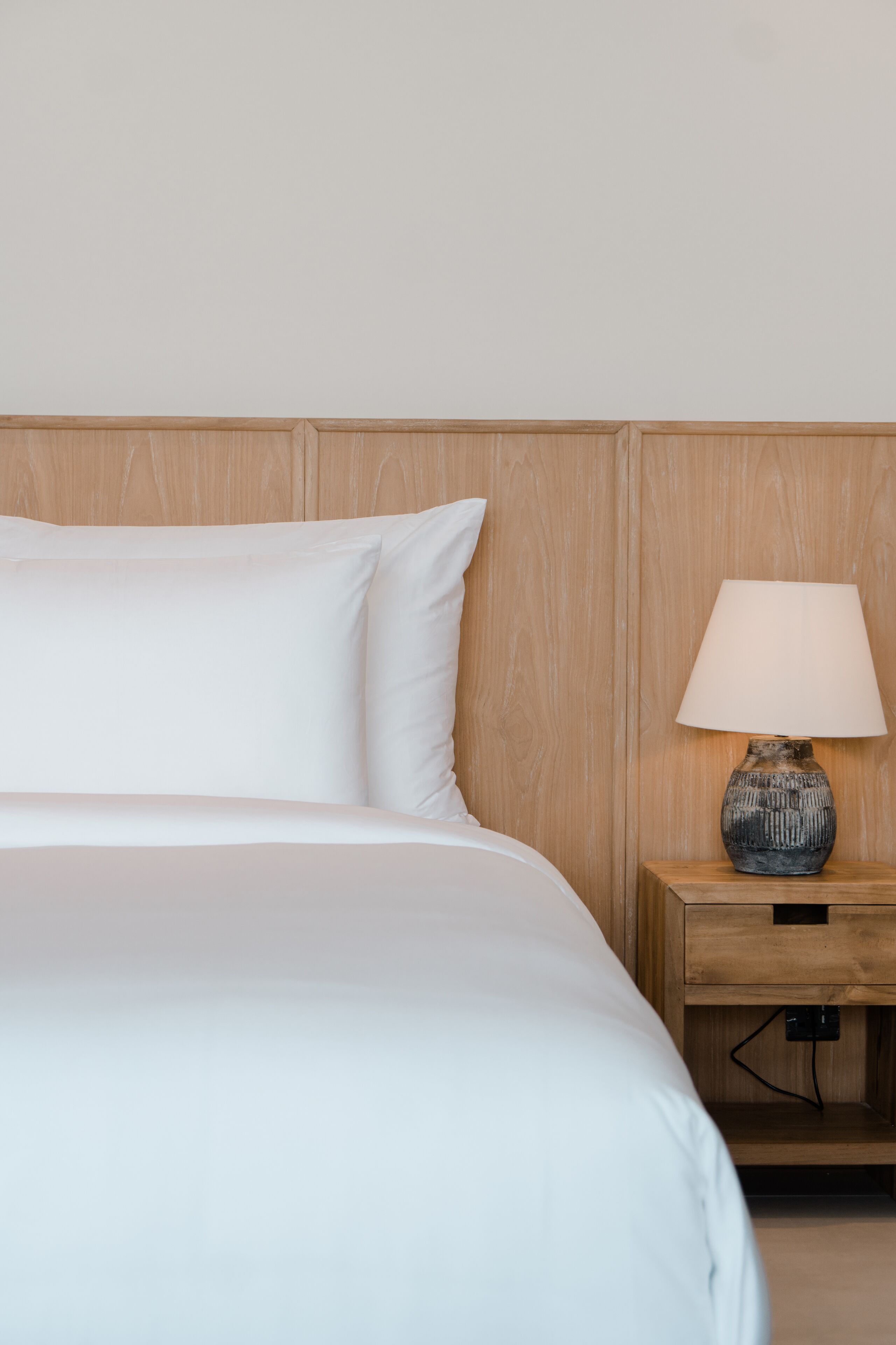 A neatly made bed with white linens and pillows, a wooden nightstand with a lamp, and a plain wall.