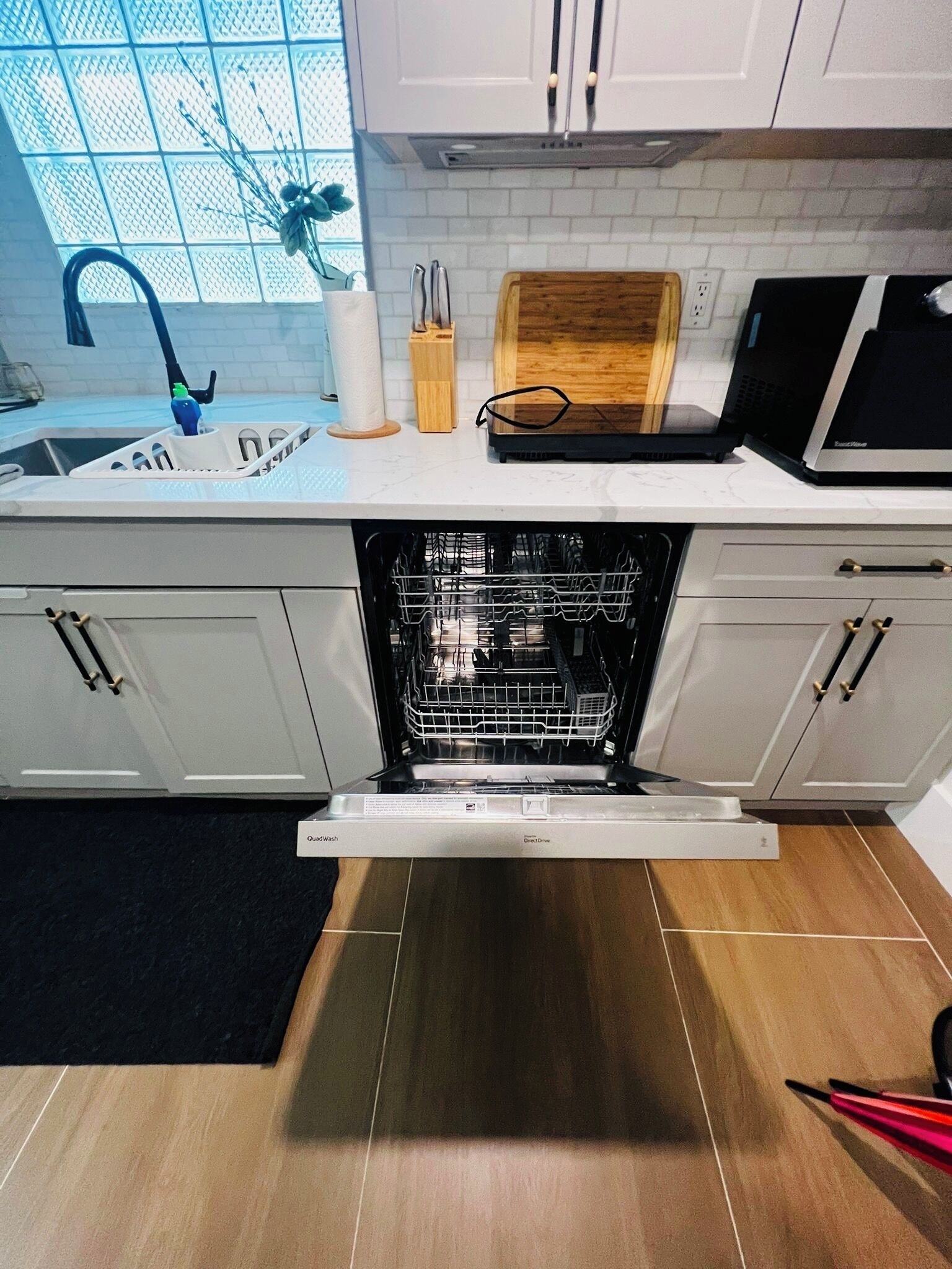 A kitchen with a dishwasher, white cabinets, a wooden cutting board, and a window with a view of the sky.