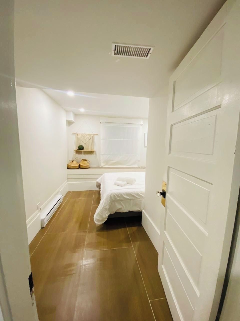 A narrow hallway leading to a bedroom with a bed, a small window, and a radiator.