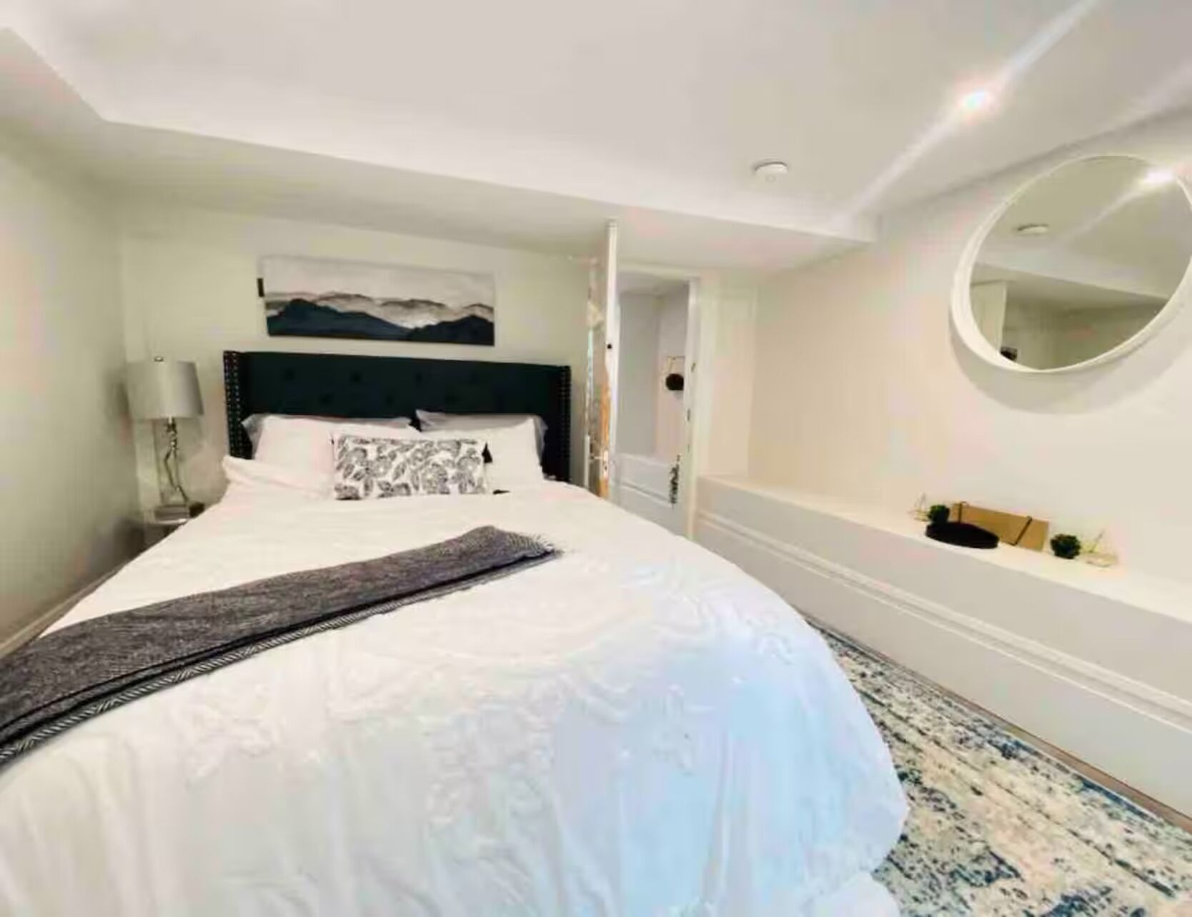 A hotel room with a large bed, a headboard, a bedside table with a lamp, a small shelf with decorative items, and a round mirror on the wall.