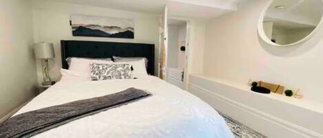 A hotel room with a large bed, a headboard, a bedside table with a lamp, a small shelf with decorative items, and a round mirror on the wall.