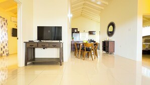 A modern living room with a flat-screen TV on a wooden console table, a dining area with wooden chairs, and a kitchen in the background.