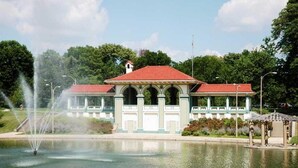 Een gebouw met een rood dak en zuilen, een fontein en een kiosk aan een waterpartij.