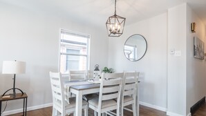 Een eethoek met een houten tafel en witte stoelen, een hanglamp, een ronde spiegel en een raam met jaloezieën.