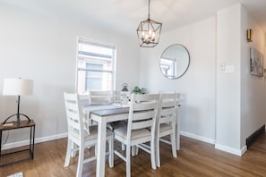 A dining area with a wooden table and white chairs, a hanging light fixture, a round mirror, and a window with blinds.