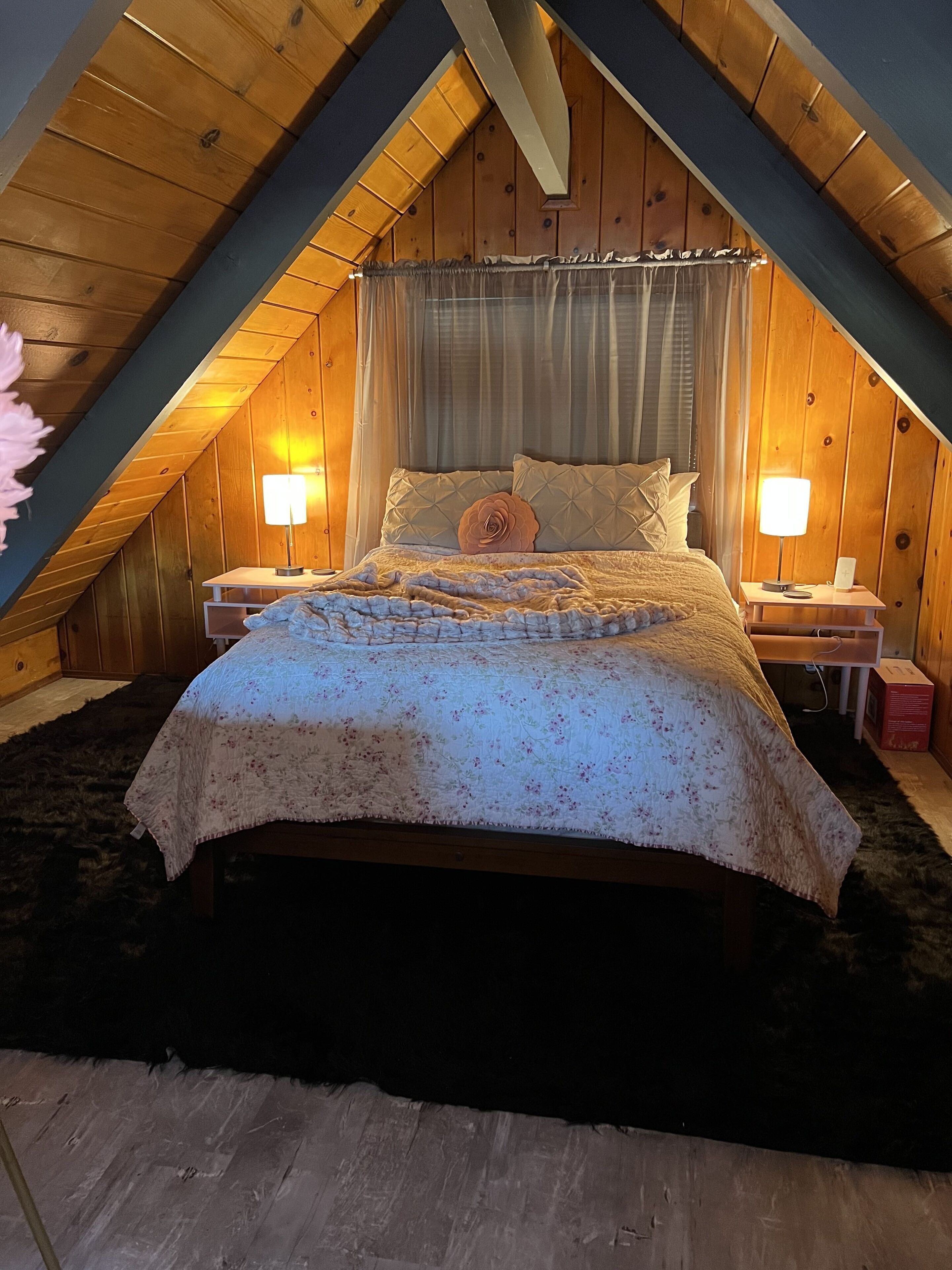A cozy attic bedroom with a sloped ceiling, a bed with a floral bedspread, two bedside lamps, and a wooden headboard.