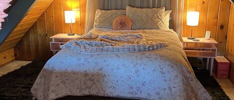 Un dormitorio acogedor en el ático, con techo inclinado, una cama con colcha estampada, dos lámparas de noche y un cabecero de madera.